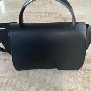 Svart handväska från ATP Atelier Arezzo Leather Handbag - Stilren svart handväska från ATP Atelier i slätt skinn med snygga guldiga metalldetaljer. Väskan har ett unikt lock med mjuka former och ett stadigt handtag. Perfekt för dig som gillar minimalistisk och modern design.Axelremsväska från ATP Atelier Försluts med lock och magnetknapp med lås Ett handtag upptill samt avtagbar axelrem  Bredd: 34 cm Höjd: 18 cm Djup: 11 cm