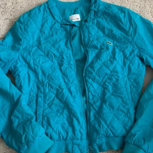 Turkos quiltad bomberjacka Lacoste - Quiltad turkos bomberjacka från Lacoste med klassisk croco-logo på bröstet. Jackan har dragkedja framtill, ribbade muddar och rund hals. Snyggt mönster i rutigt quiltat tyg och sportig vibe. Perfekt för dig som vill sticka ut med färg och stil. Den är i barnstorlek så den sitter som en xs. 