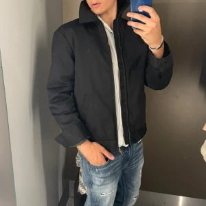 Svart bomberjacka med dragkedja - Snygg svart bomberjacka med klassisk krage och rak passform. Jackan har en hel dragkedja framtill och två sidofickor. Materialet är slätt och känns som polyester, perfekt för dig som gillar stilrena och enkla plagg. Passar till jeans och t-shirt för en chill look.