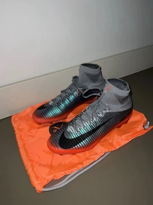 Nike fotbollsskor med högt skaft - Säljer ett par grå och svarta Nike fotbollsskor med högt skaft och snörning. Skorna har en snygg metallicblå ton och orange sula, samt en stor svart swoosh på sidan. Perfekta för dig som vill ha grymt grepp och stil på planen.