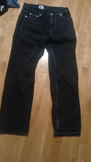 Svarta jeans från HG District - Svarta jeans med raka ben och kontrastsömmar. Bakfickan har ett broderat HG District-tryck och märket syns tydligt på linningen. Klassisk femficksmodell i jeansmaterial, perfekt för dig som gillar streetwear och vill sticka ut lite extra. Bra skick förutom att lapen med märket över bakfickan gått sönder som man även ser på en av bilderna.