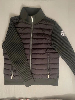 Svart cardigan från Canada Goose - Säljer en svart pufferjacka från Canada Goose i storlek M. Jackan har ribbade stickade ärmar, quiltad kropp och två fickor med dragkedja. Märkets logga sitter på ena ärmen och dragkedjan är graverad med märket. Fler bilder finns i DM. Priset kan diskuteras.