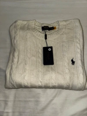 Vit kabelstickad tröja Polo Ralph Lauren - Säljer en vit kabelstickad tröja från Polo Ralph Lauren i storlek M. Tröjan har rund hals, ribbade muddar och det klassiska broderade logot på bröstet. Materialet är en mix av ull, akryl och nylon för extra komfort och värme.