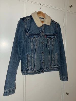 Levi's jeansjacka med teddykrage M - Säljer en klassisk original blå jeansjacka från Levi's med vit teddykrage. Jackan har bruna knappar, två bröstfickor med lock och sidofickor. Insidan är fodrad med mjuk teddy för extra värme. Perfekt för dig som vill ha en chill och tidlös look.
