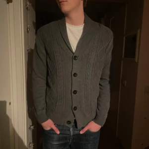 Grå kofta från Selected Jeans - Säljer en snygg grå kofta från Selected Jeans i storlek M. Koftan har kabelstickat mönster, ribbade muddar och fem svarta knappar framtill. Perfekt för lager-på-lager och chill stil. Materialet är mjuk bomull och polyester.