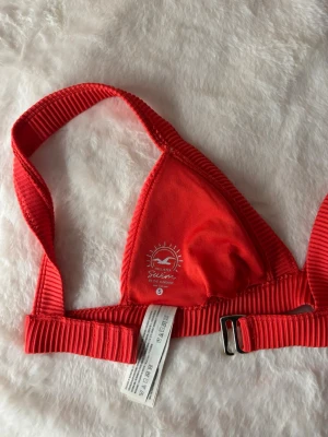 Röd ribbad bikiniöverdel från Hollister - Röd bikiniöverdel från Hollister med ribbad struktur och trekantsformade kupor. Justerbart band med metallspänne baktill. Storlek S.