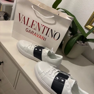 Valentino Garavani vita sneakers - Snygga vita sneakers från Valentino Garavani med bred svart läderdetalj över sidan och coola nitar på hälen. Skorna har snörning och rund tå, samt diskret logga på plösen och sulan. Perfekta för dig som vill ha en lyxig och stilren look.