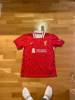 Röd Liverpool fotbollströja Nike - Säljer en röd Liverpool fotbollströja från Nike med vita och gula detaljer. Tröjan har tunna gula ränder och klubbmärke på bröstet, samt sponsortryck. Perfekt för dig som älskar fotboll och vill visa ditt stöd för Liverpool FC.
