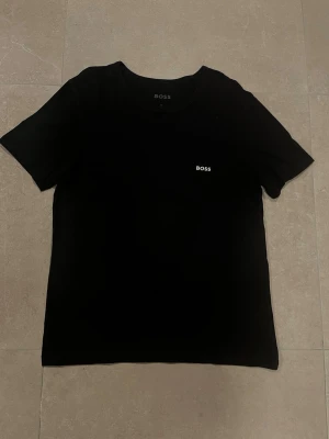 Svart BOSS t-shirt med logga - Svart t-shirt från BOSS i mjuk bomull med klassisk rund hals och korta ärmar. Diskret BOSS-logga i vitt på bröstet ger en clean och stilren vibe. Perfekt för dig som gillar enkel och snygg design.