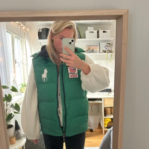 Grön dunväst från Polo Ralph Lauren - Grön dunväst från Polo Ralph Lauren med stor vit logga på bröstet. Västen har huva och dragkedja. Fint skick!