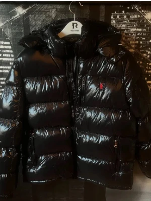 Svart pufferjacka från Polo Ralph Lauren - Säljer en svart, glansig pufferjacka från Polo Ralph Lauren med röd broderad logga på bröstet. Perfekt för kalla dagar och riktigt snygg streetstil. Säljer den då den var för stror för mig