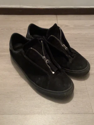 Axel arigato skor  - Svarta sneakers från Arigato i mocka med coola dragkedjor framtill istället för klassisk snörning. Snygg platt sula och rund tå, samt detaljer i svart skinn vid hälen. Perfekt för dig som gillar stilrena och unika skor.