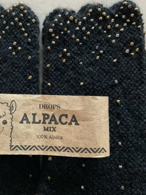 Svart stickad alpaka-väst med pärlor - Unika fingerlösa vantar, 100 % alpackaull, helt handgjorda med förstklassigt hantverk. Glittriga små pärlor är invirade för hand över hela plagget, och den vågiga kanten ger en originell och iögonfallande form. Perfekt för dig som vill ha något extra till din outfit.