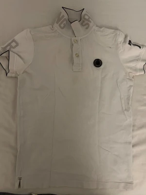 Vit polotopp Sail Racing med tryck - Vit polotopp från Sail Racing med korta ärmar och knappar framtill. Kragen har grått tryck med bokstäver och svart kant. Toppens material är bomull och har en rund svart logotyp på bröstet. Snygg och sportig design som passar till många stilar.