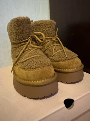 Bruna moon boots från Graceland - Säljer ett par bruna moon boots från Graceland med fluffig pälsimitation och mockadetaljer. Skorna har snörning runt hela skaftet och en rejäl platt sula som ger extra höjd. Perfekta för kalla dagar med sin mysiga look och chunky design.