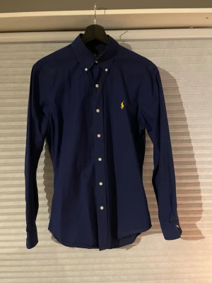 Marinblå skjorta från Ralph Lauren - Snygg marinblå skjorta från Ralph Lauren i slim fit med klassisk krage och knappar framtill. Den är i bra skick och säljs då den blivit liten. Pris 299kr. //David