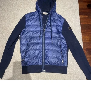 Blå Moncler Cardigan - Unik och stilren cardigan jacka från Moncler med quiltad pufferfront och stickade ärmar. Jackan har huva med snörning, två fickor med dragkedja och ribbad nederkant. Passar dig runt 170-180 ge och ta