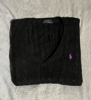 Svart stickad tröja från Polo Ralph Lauren - Svart stickad tröja från Polo Ralph Lauren med kabelstickat mönster och lila broderad logga på bröstet. Tröjan har v-ringad hals och är perfekt för dig som gillar klassisk stil med en twist. Passar dig som vill ha en snygg och enkel look.