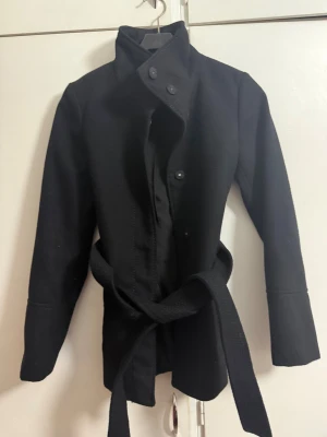 Svart kappa med bälte från H&M - Svart kappa från H&M i storlek 34 med högt krage, knappar framtill och ett snyggt bälte i midjan. Jackan har långärmade ärmar och en stilren, rak passform som ger en clean look. Perfekt för dig som gillar minimalistisk stil och vill ha något som funkar till många outfits.