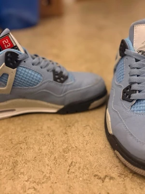 Nike Air Jordan 4 University Blue - Nike Air Jordan 4 University Blue sneakers med ljusblå mocka och mesh, svarta och vita detaljer samt speckled midsole. Ikonisk Jumpman-logga på tungan och sulan. Skorna har snörning och platt sula, samt en rund tå. Kommer med originalkartong.