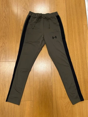 Under Armour träningsbyxor med svarta ränder - Snygga olivgröna träningsbyxor aldriganvända från Under Armour i storlek S. Byxorna har svarta sidopaneler, snörning i midjan och är gjorda i ett mjukt syntetmaterial som andas. Perfekta för gymmet eller sportaktiviteter. Fitted passform för en modern look.