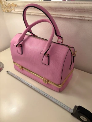 Rosa handväska med gulddetaljer - Säljer en snygg rosa handväska med guldfärgade metalldetaljer och praktiskt fack i botten. Väskan har två stadiga handtag och en dragkedja upptill. Materialet är syntet med en diskret ribbad struktur. Perfekt för dig som gillar färg och vill ha plats för allt viktigt.