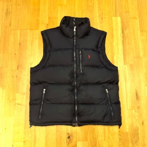Svart dunväst från Polo Ralph Lauren - Snygg svart dunväst från Polo Ralph Lauren med hög krage och tre dragkedjefickor. Klassisk design med röd broderad logga på bröstet. Perfekt att slänga över en hoodie för extra värme. Stängs med dragkedja framtill.