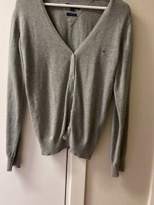 Grå kofta från Tommy Hilfiger - Grå kofta från Tommy Hilfiger med v-ringning och små knappar framtill. Diskret logga broderad på bröstet. Långärmad och tillverkad i mjuk bomull, perfekt för lager-på-lager. Ribbstickade muddar vid ärmslut och nederkant ger en snygg passform.