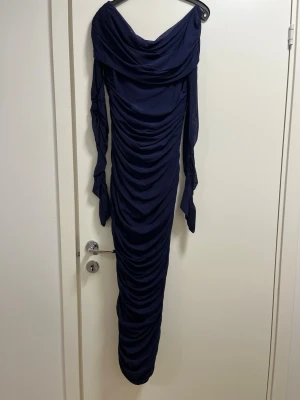 Marinblå långklänning med drapering - Säljer en marinblå långklänning med snygg draperad design och långa ärmar. Klänningen har en bred, nedhasad krage och är figurnära med rynkade detaljer längs hela längden. Perfekt för dig som vill ha en elegant och trendig look.