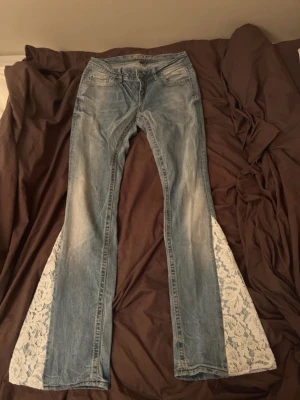 Bootcut jeans med spets - Unika ljusblå bootcut jeans från Fishbone med breda ben och insydd vit spets i blommigt mönster längs utsidan av benen. Snygga fickor med detaljer, skulle säga att dem passar storlek xs kontakta vid frågor och funderingar