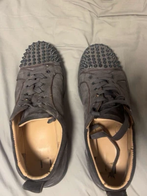 Grå sneakers med nitar på tån - Säljer ett par grå sneakers med coola nitar över hela tån. Skorna har snörning och är gjorda i mocka med en rund tå och platt sula. Insidan är beige och de har en urban vibe som sticker ut. Perfekta för dig som gillar att synas och vill ha något unikt på fötterna.