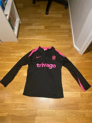 Chelsea Tracksuit 24/25 - Säljer ett  träningsset från Nike med Chelsea FC-design, säsong 24/25. Setet består av en svart zip-träningströja och matchande svarta träningsbyxor, båda med rosa detaljer och klubbens lejonemblem. Tröjan har “trivago” tryckt framtill och rosa ränder längs axlar och ärmar. Byxorna har elastisk midja och rosa Nike swoosh.