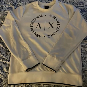 Vit sweatshirt från Armani Exchange - Snygg vit sweatshirt från Armani Exchange i storlek M. Tröjan har ett stort svart tryck med A|X-logga på bröstet och svarta detaljer vid muddar och nederkant. Rund hals och långärmad modell, perfekt för en clean och stilren look. Skriv vid frågor eller bud