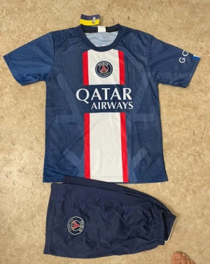 PSG Mbappé matchställ blå - Säljer ett PSG matchställ med Mbappé 7 på ryggen. Tröjan är marinblå med vita och röda detaljer, Qatar Airways-logga och PSG-emblem. Tillhörande shorts i samma färg med PSG-logga. Perfekt för dig som älskar fotboll och vill bära ditt favoritlag.