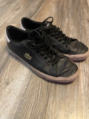 Svarta sneakers från Lacoste - Säljer ett par svarta sneakers från Lacoste med klassisk krokodil-logga på sidan. Skorna har snörning, vit sula och detaljer i vitt baktill. Tillverkade i skinn för en clean och stilren look som funkar till det mesta.