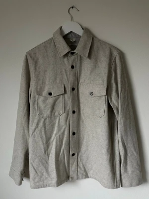 Ljusgrå overshirt från ARKET i ullmix - Snygg ljusgrå overshirt från ARKET i ullblandad twill. Regular fit med klassisk krage, två stora bröstfickor med knappar och svarta knappdetaljer framtill och vid ärmslut. Perfekt lager-på-lager-plagg för dig som gillar stilren och enkel design. 5L18