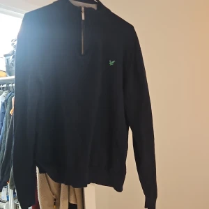 Marinblå half zip tröja Lyle & Scott - Snygg marinblå långärmad tröja från Lyle & Scott med half zip och ribbad krage i grått. Broderad grön logga på bröstet. Perfekt för dig som gillar stilrena och klassiska plagg med en sportig touch.