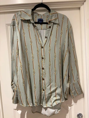 Grön mönstrad skjorta från GANT - Snygg grön skjorta från GANT med unikt mönster av nycklar och ränder i guld och brunt. Skjortan har klassisk krage, knappar framtill och långa ärmar med manschett. Materialet känns mjukt och har en lätt glansig finish, perfekt för dig som gillar detaljer.