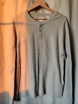Grå långärmad tröja från Levi's - Säljer en grå långärmad tröja från Levi's i storlek L. Tröjan har rund hals, tre knappar framtill och är tillverkad i mjuk bomull. Perfekt för chill dagar och enkel att matcha med jeans eller joggers.