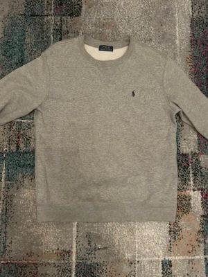 Grå sweatshirt från Polo Ralph Lauren - Klassisk grå sweatshirt från Polo Ralph Lauren med rund halsringning och den ikoniska broderade loggan på bröstet. Tröjan har långa ärmar och är tillverkad i mjuk bomull för maximal komfort. Perfekt till jeans eller sweatpants.