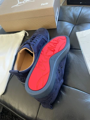 Mörkblå sneakers från Christian Louboutin - Snygga mörkblå sneakers från Christian Louboutin med lyxig röd sula och diskret logga. Skorna är tillverkade i mocka med rund tå och snörning. Insidan är beige och sulan har en cool signaturdetalj. Perfekt för dig som vill sticka ut med stil.