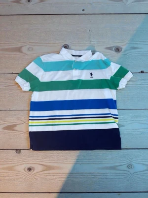 Randig pikétröja från U.S. Polo Assn XL - Fräsch pikétröja från U.S. Polo Assn i storlek XL. Tröjan har breda ränder i vitt, grönt, turkos, blått, gult och mörkblått. Klassisk krage med två knappar och broderad USPA-logga på bröstet. Perfekt för dig som gillar färg och sportig stil. 