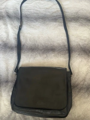 Svart axelväska  - Säljer en stilren svart axelväska med klassisk fyrkantig form och lång justerbar rem. Insidan har flera fack, inklusive ett med dragkedja, och är fodrad med rutigt tyg. Perfekt för dig som vill ha ordning på grejerna och gillar enkel design.🌸 Sparsamt använd