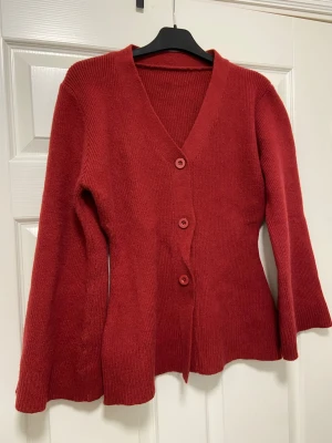 Röd stickad kofta med v-ringning - Helt ny och oanvänd kofta. Koftan har långa ärmar med lätt utsvängd form och ribbade muddar. Figursydd i midjan. Perfekt att slänga på sig när det är lite kyligt ute och passar perfekt till jul. Materialet är mjukt och ger en skön känsla. Superfin!