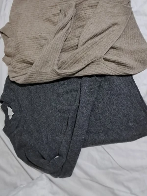 Grå och beige stickade koftor - Två mysiga stickade koftor i storlek M. Den ena är mörkgrå med ribbad struktur och rund hals, den andra är beige med bred krage och ribbad design. Perfekta för lager-på-lager och chill dagar. Snygga att matcha med jeans eller leggings.