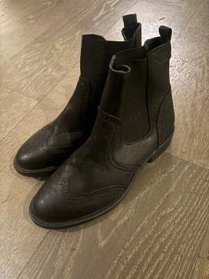 Svarta chelsea boots i skinn - Säljer ett par svarta chelsea boots i skinn med klassisk rund tå och dekorativa broguemönster. Bootsens elastiska paneler på sidorna gör dem enkla att ta på och av. Perfekta för dig som gillar stilrena och tidlösa skor med snygga detaljer.