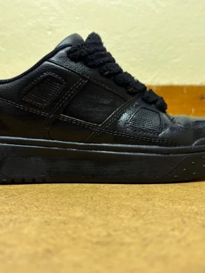 Svarta sneakers från Zara med grova snören - Säljer ett par svarta sneakers från Zara med grova svarta snören och detaljerad sömnad. Skorna har en rund tå, platt sula med rutmönster undertill och är tillverkade i skinnliknande syntetmaterial. Perfekt för dig som gillar chunky stil och svart på svart storlek 40.