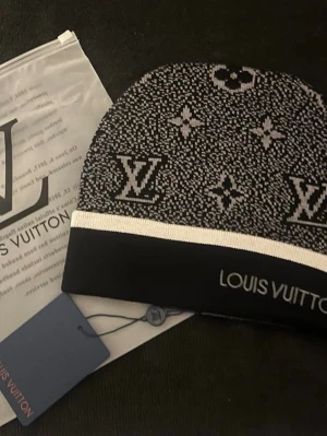 Svart mössa med vita detaljer  - Säljer en snygg svart och vit mössa med klassiskt monogrammönster och logotyp framtill. Mössan har en bred vit kant och är stickad i mjukt material. Perfekt för dig som vill ha en lyxig accessoar till vintern.