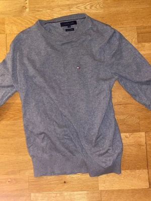 Grå tröja från Tommy Hilfiger S - Snygg grå tröja från Tommy Hilfiger i storlek S. Tillverkad i en mjuk mix av bomull och kashmir, med diskret logga broderad på bröstet. Rund hals och ribbade muddar vid ärmslut och nederkant. Perfekt för dig som gillar stilrena och klassiska plagg.