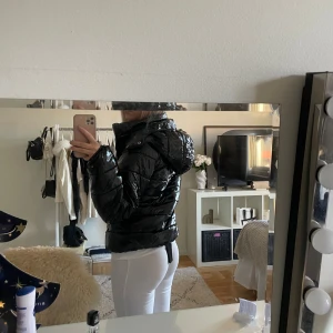 Svart glansig pufferjacka med huva - Säljer en svart glansig pufferjacka med huva och dragkedja framtill. Jackan har en quiltad look och är lite croppad i modellen, perfekt för dig som gillar streetstyle. Passformen är loose och jackan har långa ärmar. Snygg att styla med jeans eller leggings.